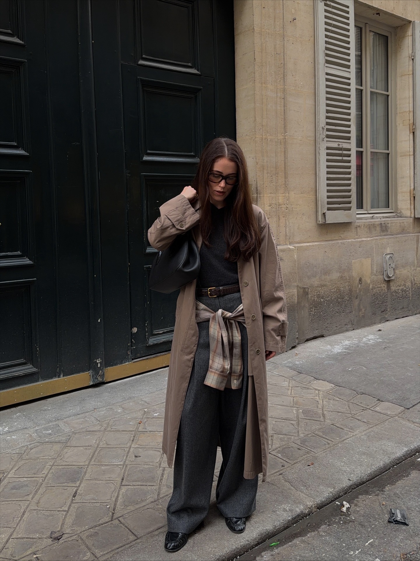Annabel wears a beige trench coat, black top, grey tailored trousers and checked shirt