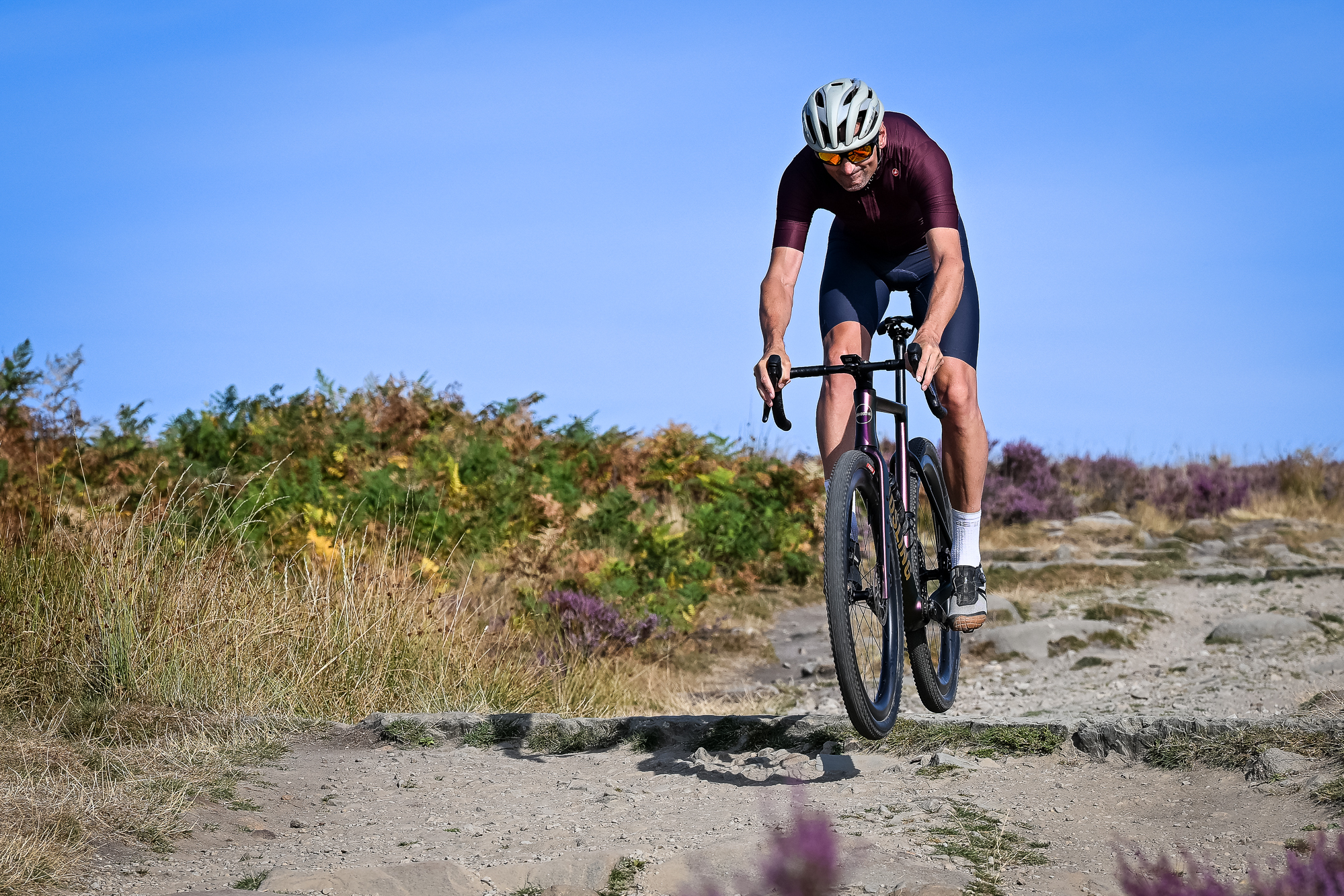 Man jumping a purple gravel bike on a sandy trail wearing grey shoes, blue shorts, a purple top and a beige helmet.