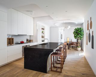 A kitchen with a black marble island, wooden chairs, white storage shelving and a potted plant.