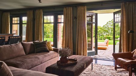 Large living room area with L-shaped sofa, padded coffee table and rug. Double doors are open revealing bright outdoor area with outdoor seating, partially covered by yellow curtains.