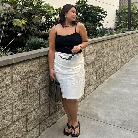 Marina wears black tank top, white linen skirt, and black flip flops while posing for a picture. 