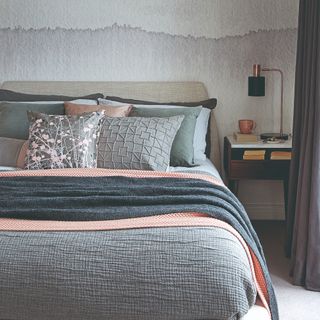King sized bed with cool grey, silver and blue bedding with a hint of pink, next to bedside table and navy curtains