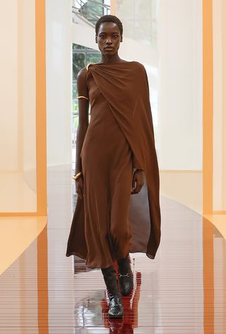 one of the best chunky cuff bracelets shown in a photo from Gucci's spring 2025 runway show with a model wearing a brown cape gown with black boots with horse bit hardware and layered gold cuffs