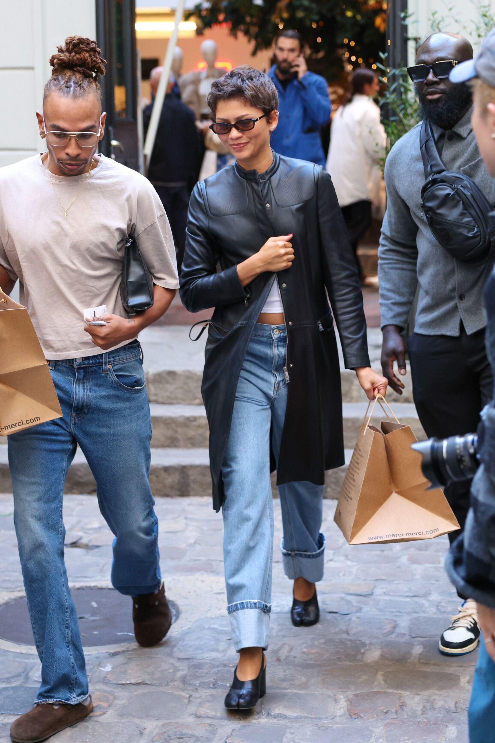 Zendaya steps outside in Paris wearing a black leather coat with blue cuffed jeans, heeled Tabis and black sunglasses. She carries a paper shopping bag.