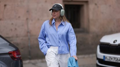 Sonia Lyson wearing Zara hose, Storets blue and white striped long sleeve buttoned cropped flannel, Bottega Veneta blue leather pouch bag, Axel Arigato dark grey logo cap, Apple AirPods Max olive color headphones on June 28, 2023 in Berlin, Germany.