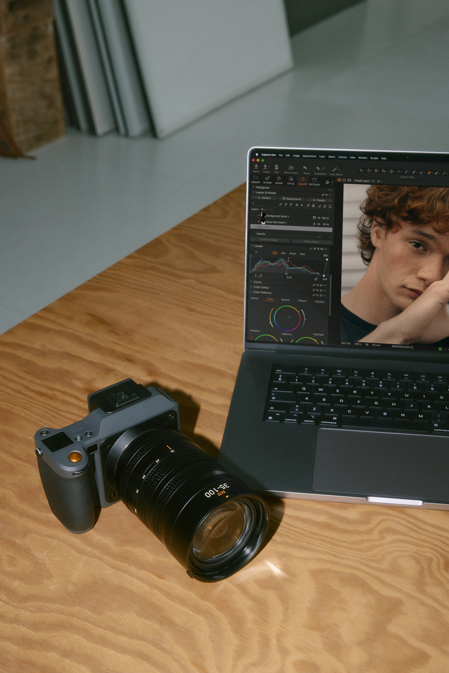 Camera with 35-100mm lens beside a laptop displaying photo editing software and a portrait of a person on a wooden table