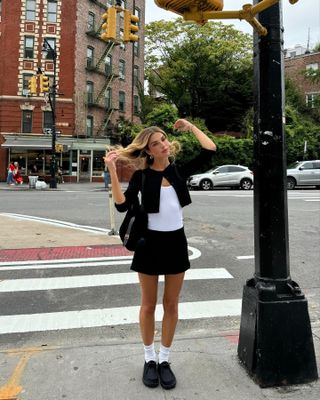 Kit wears a black cropped cardigan, white tank top, black skirt, white socks, a black shoulder bag, and black flats.