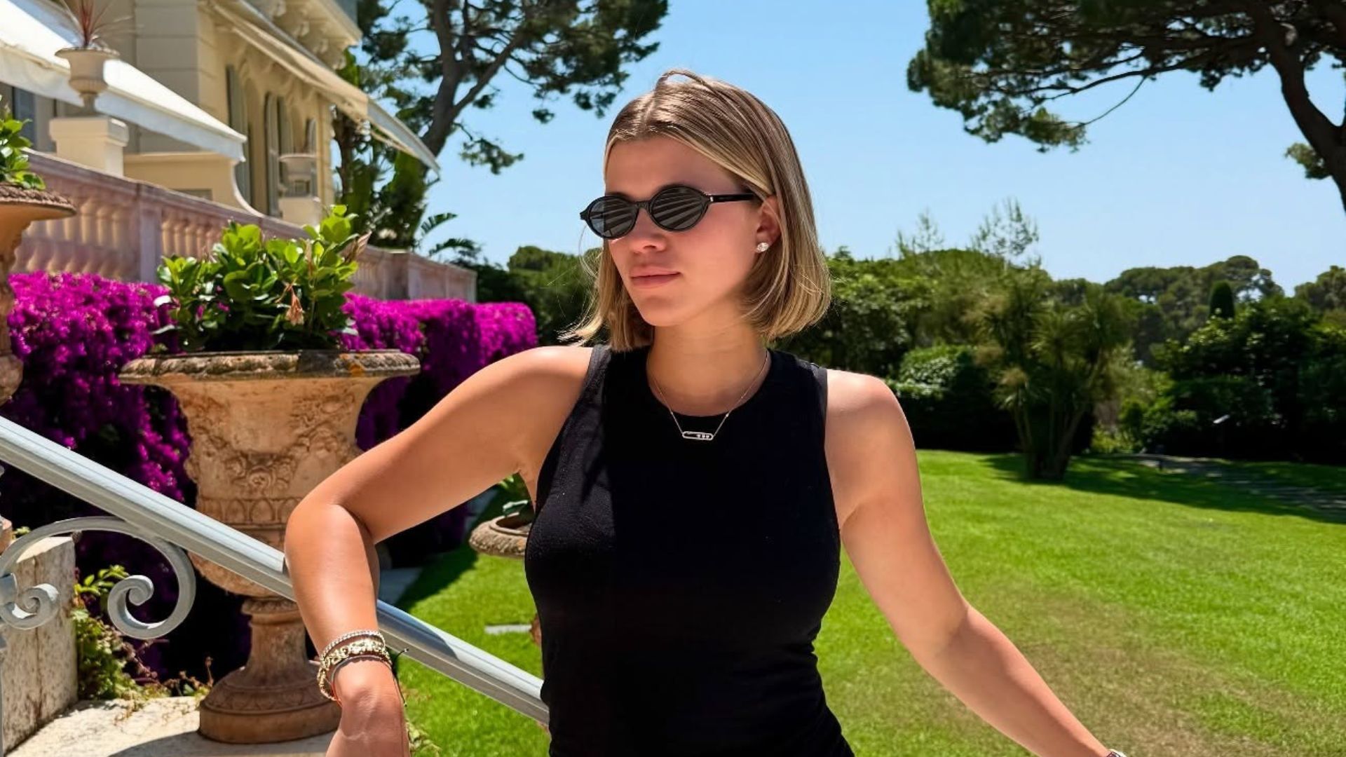 Sofia Richie sits outfit on the grass wearing black trousers with a black top and green trainers.