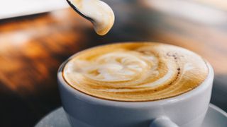 A cup of frothy coffee, using a spoon to scoop some of the froth off the top of the cup