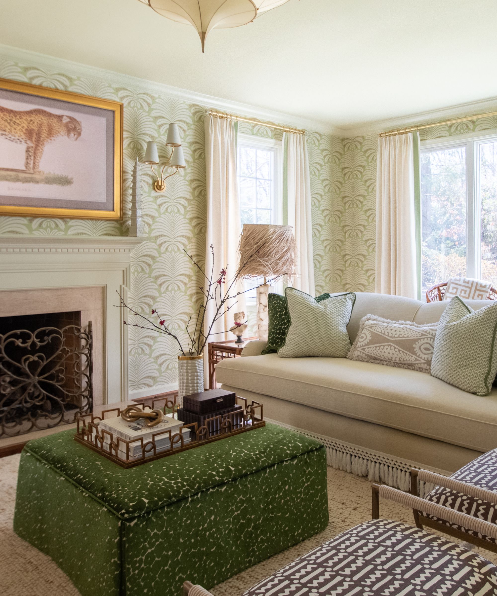 Living room with patterned pale green wallpaper, a green velour leopard print ottoman, a beige fringed sofa, two upholstered chairs, and a fireplace with a large print of a leopard above.