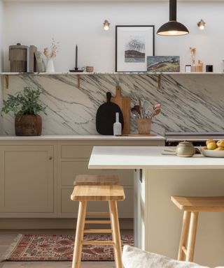 A kitchen with white kitchen island with three wooden stools, with white kitchen counters and a marble splashback behind it