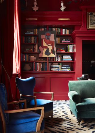 A red painted living room with a built-in bookshelf on the wall. There is a zebra print carpet, two gemstone blue accent chairs and a green velvet sofa.