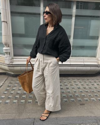 British influencer Lucy Alston poses on a London sidewalk wearing black sunglasses, a black linen shirt, camel suede tote bag, cropped and cuffed neutral linen pants, and black strappy flat sandals