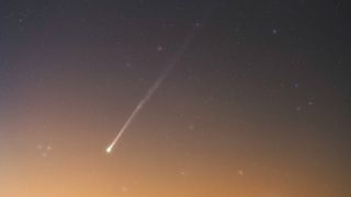 A comet is pictured shining in a field of multicolored stars as a warm yellow-orange glow suffuses the horizon.