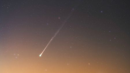 A comet is pictured shining in a field of multicolored stars as a warm yellow-orange glow suffuses the horizon.