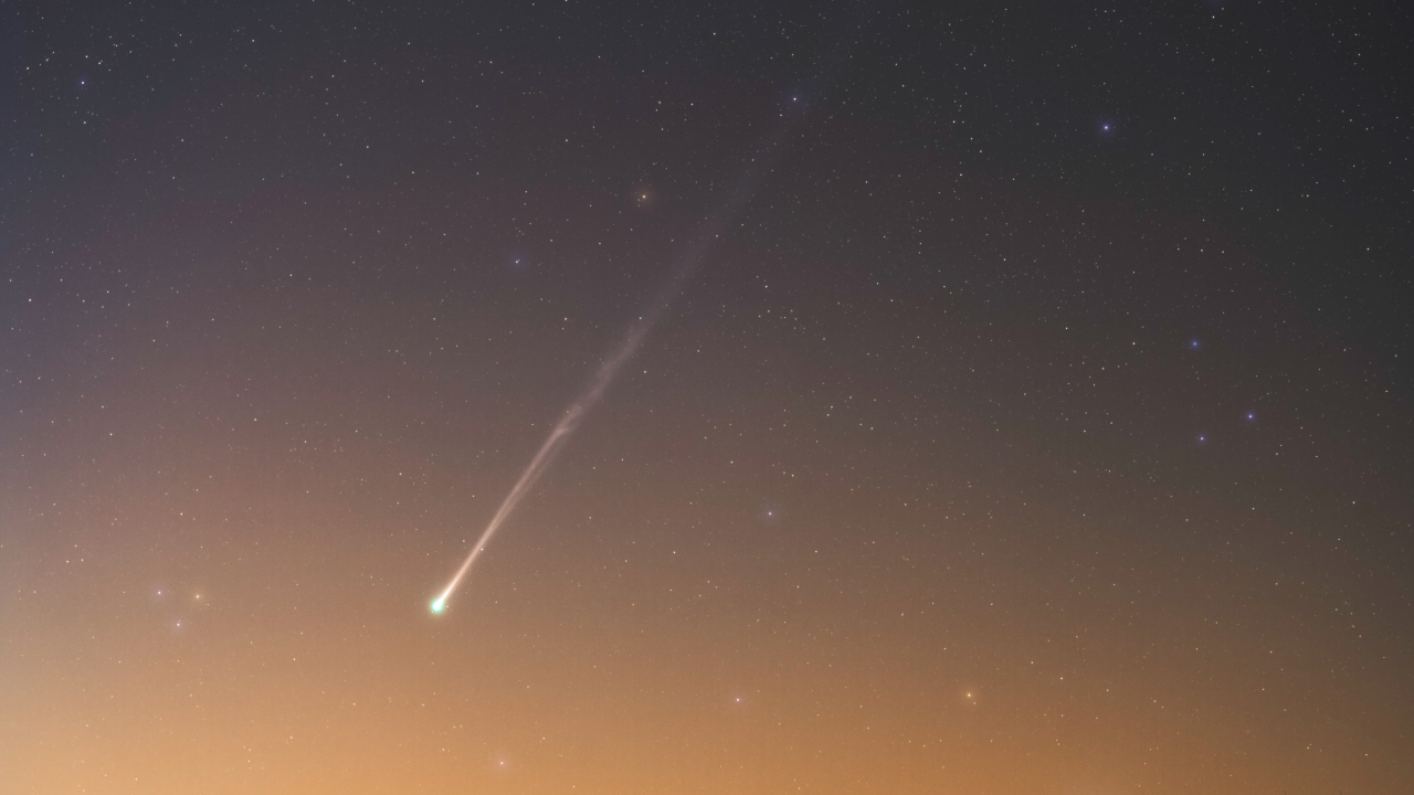 A comet is pictured shining in a field of multicolored stars as a warm yellow-orange glow suffuses the horizon.