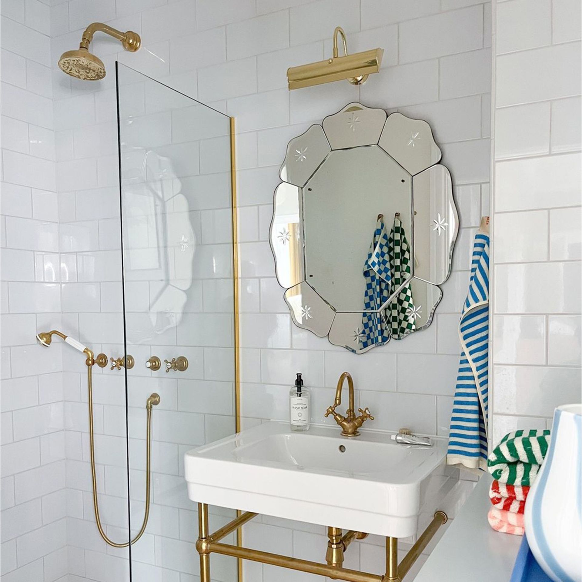 Cermin dinding bergerigi di atas wastafel di kamar mandi putih dengan perlengkapan kuningan dan handuk berwarna bergaris