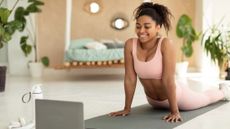 A woman practices yoga in a living room on a mat and look towards a laptop screen in front of her. She is holding cobra pose, with her legs laid straight behind her and her torso held aloft by her straightened arms. Behind her we see a couch and plants.