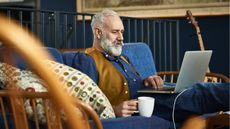 An older man sits on his sofa and looks at his laptop.