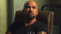 Shemar Moore is shown as Hondo on S.W.A.T. Exiles, sitting in a desk chair with a bare cement wall in the background. He's wearing a tight black shirt and looking up as he talks to an unseen recruit.