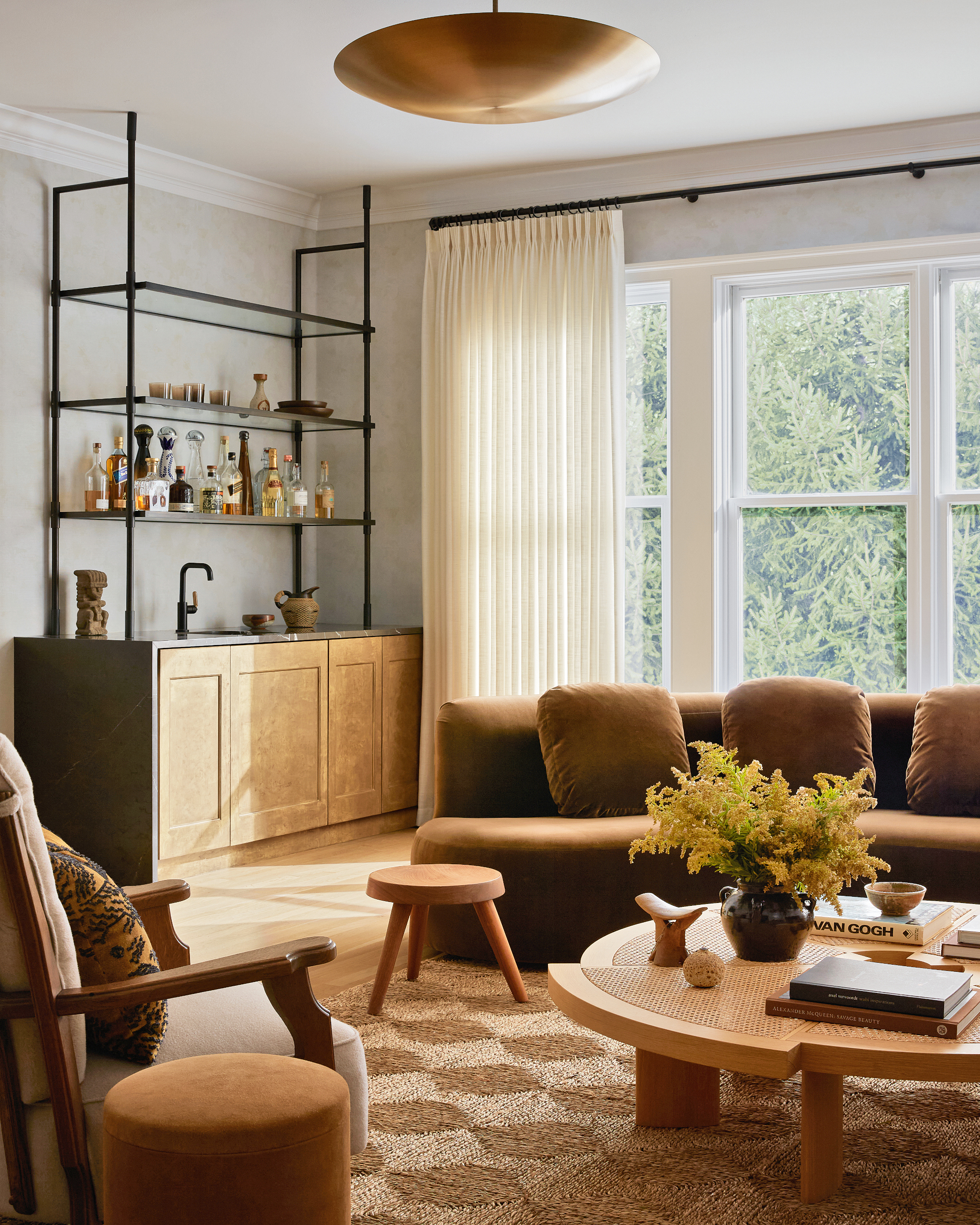 Corner of a living room with deep ochre sofa, bar area with burl wood cabinetry and grey countertop, round wood coffee table and wood stools
