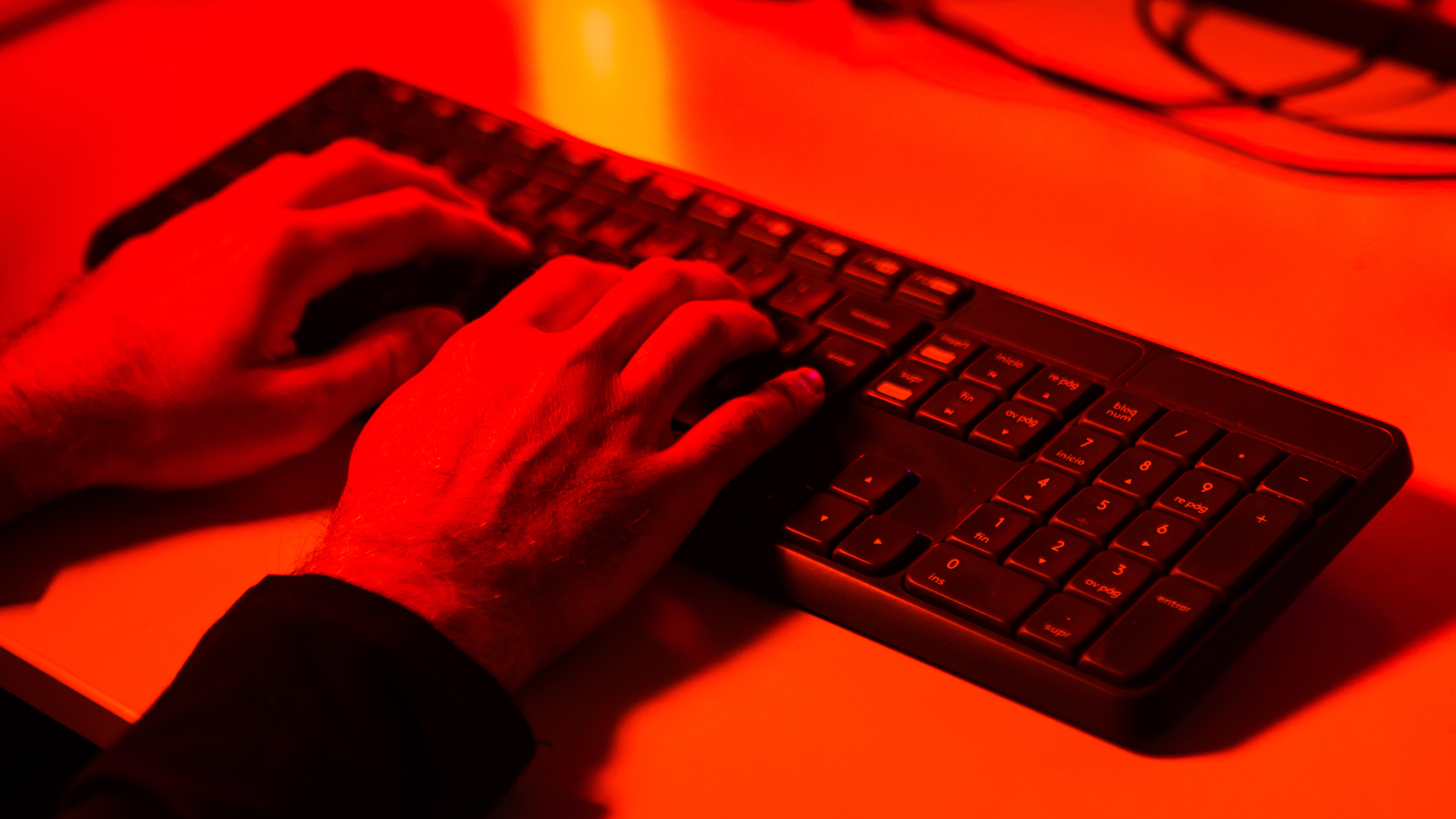 Hands of a person typing and stealing information on a computer keyboard, illuminated with red light. Concept of cybersecurity, theft, hacker, identity and crime.