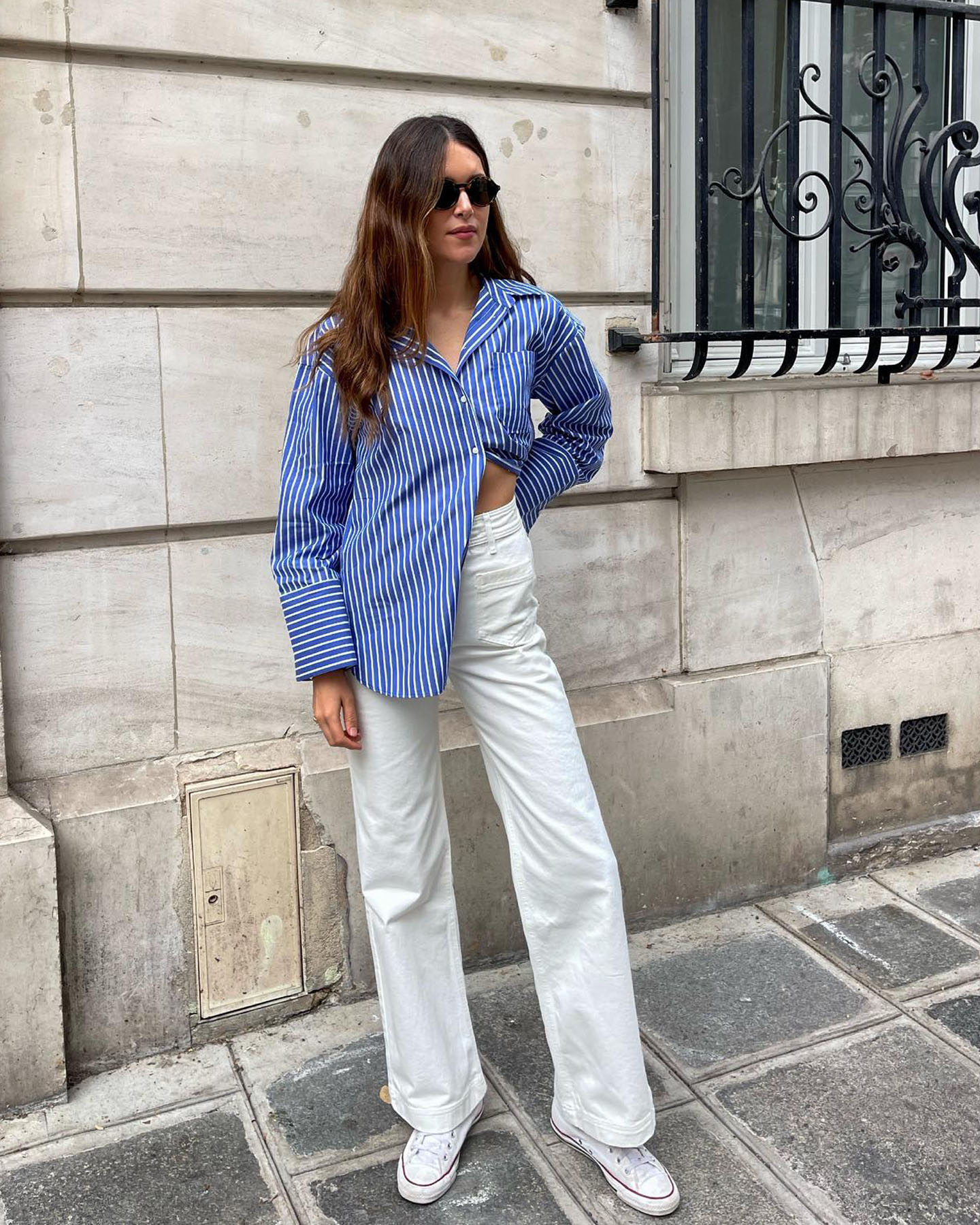 a stylish woman poses on a street in Paris wearing round sunglasses, a casual outfit with a striped blue button-down shirt, white wide-leg jeans, and white Converse sneakers