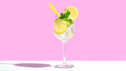 Hugo spritz in glass with ice, lemon and mint on a pink background.