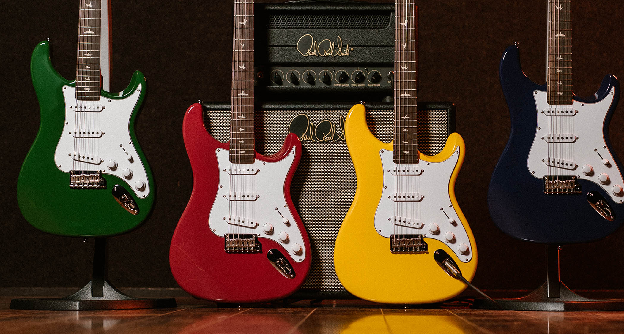 PRS SE Silver Sky 2025: refreshed for the brand&#039;s 40th anniversary, the SE Silver Sky John Mayer signature model is pictured in its new Laurel Green, Derby Red, Dandy Lion and Trad Blue finishes. A PRS tube amp and speaker cabinet is in the background.