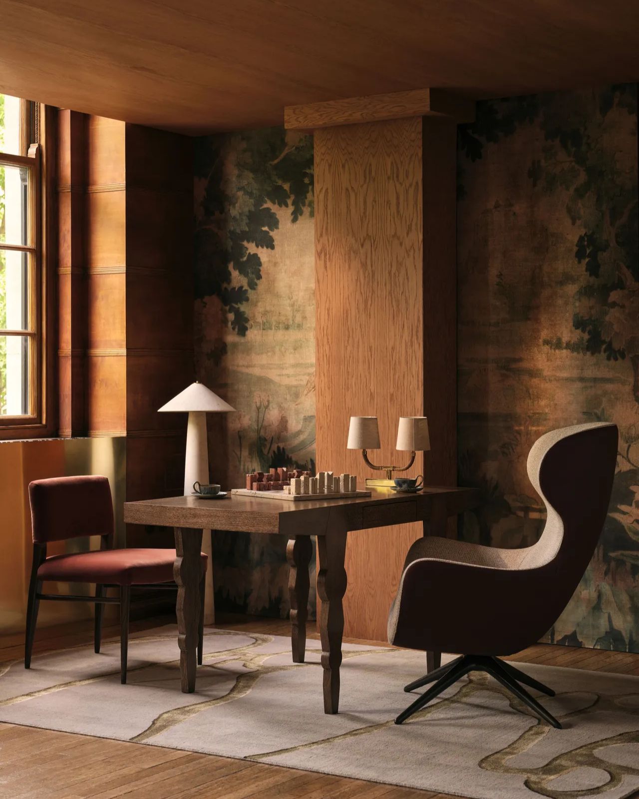 corner of room with timber column and ceiling, tapestry-style wall mural, table with chess est, lamps, and two chairs on either side, and white rug with gold squiggles through it