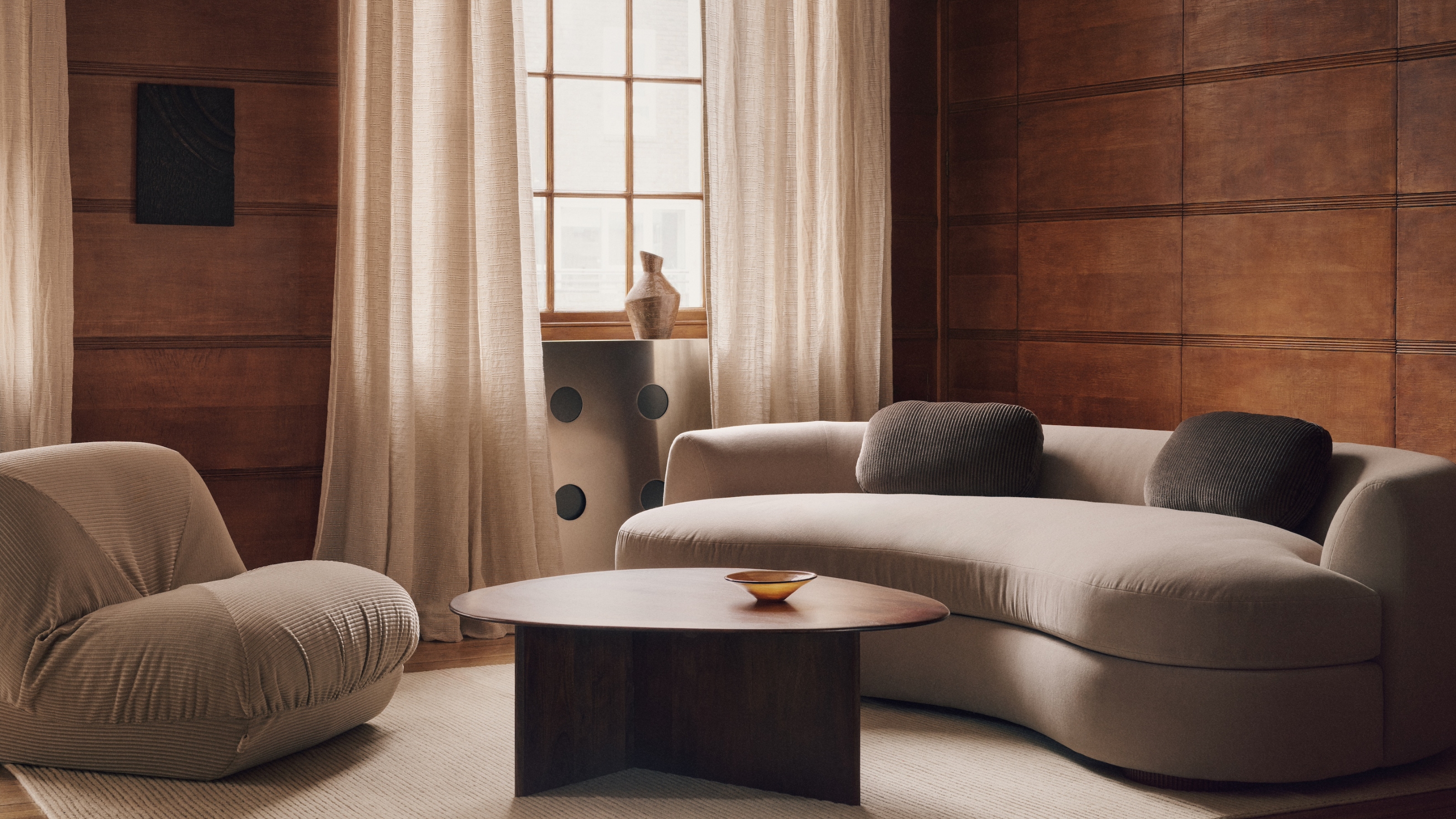 A wood-panelled living room with a curved, gray sofa and a matching bubble accent chair. There is an organically shaped coffee table in the center of the room and a window with rustic linen curtains. 
