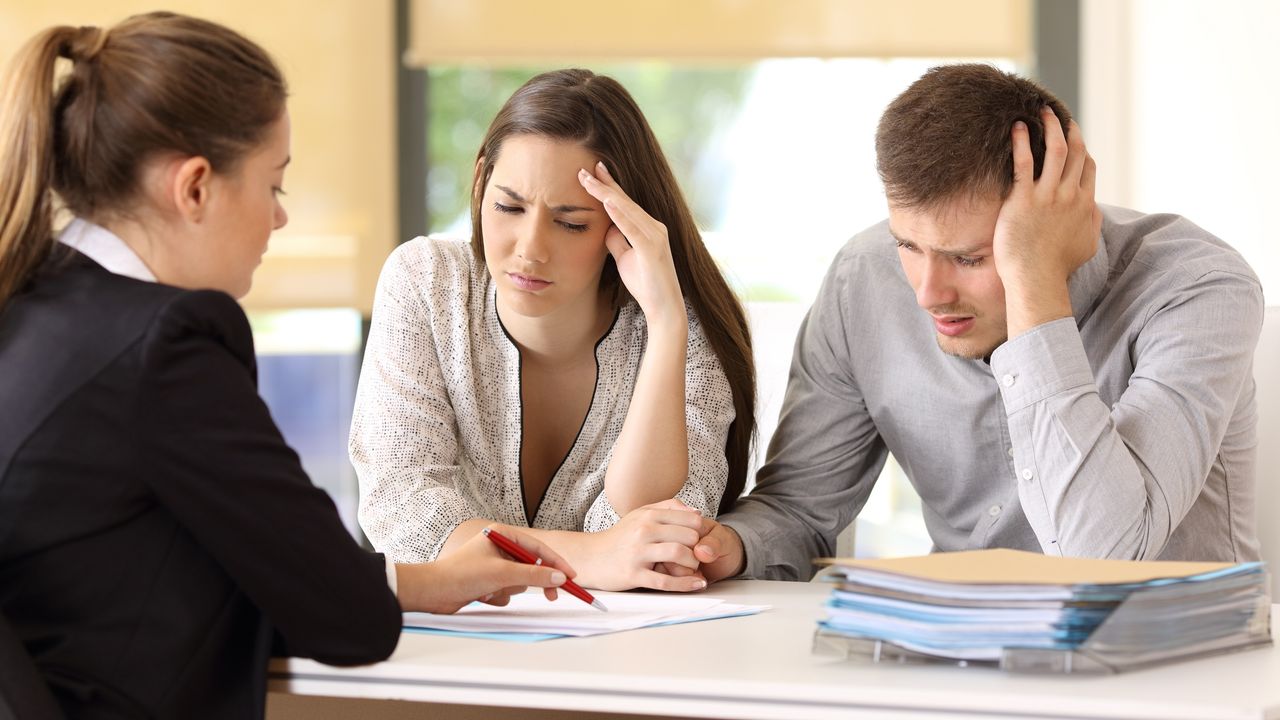 A confused-looking couple look at paperwork on a desk while a lawyer looks on.