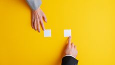 A woman's hand and a man's hand point at two separate white sticky notes against a yellow background.