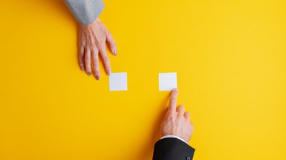 A woman's hand and a man's hand point at two separate white sticky notes against a yellow background.