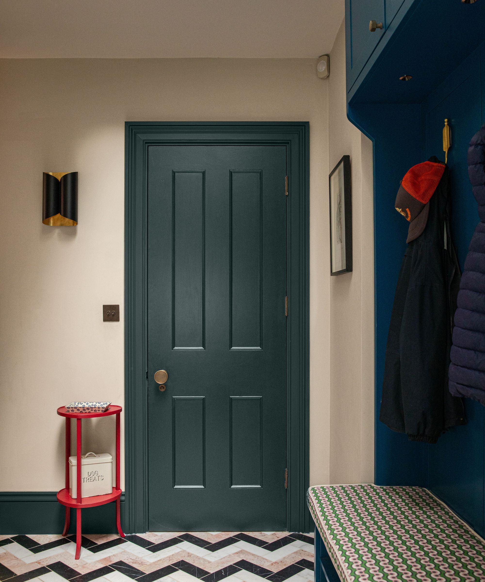 Mudroom with dark teal front door, chevron pink, black, and white tiled floor, a built-in bench and storage unit painted dark blue, and a small bright red side table by the door.