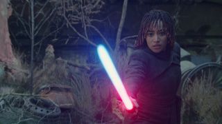 A young woman wearing dark robed is holding a lightsaber in front of her. She is changing the color of the lightsaber from blue to red.