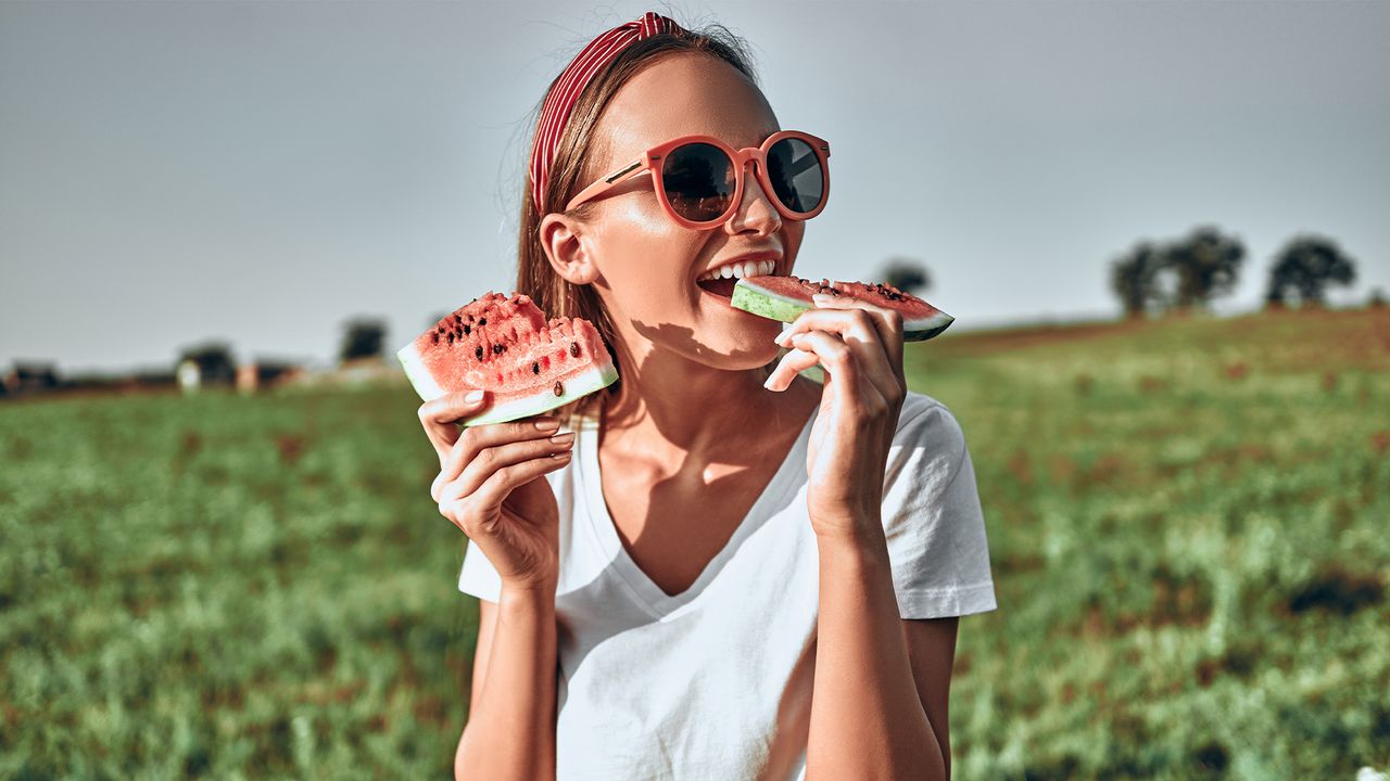 Sexy girl in red sunglasses holds a cut watermelon in her hands. Happy woman in trendy red sunglasses eat watermelon on nature