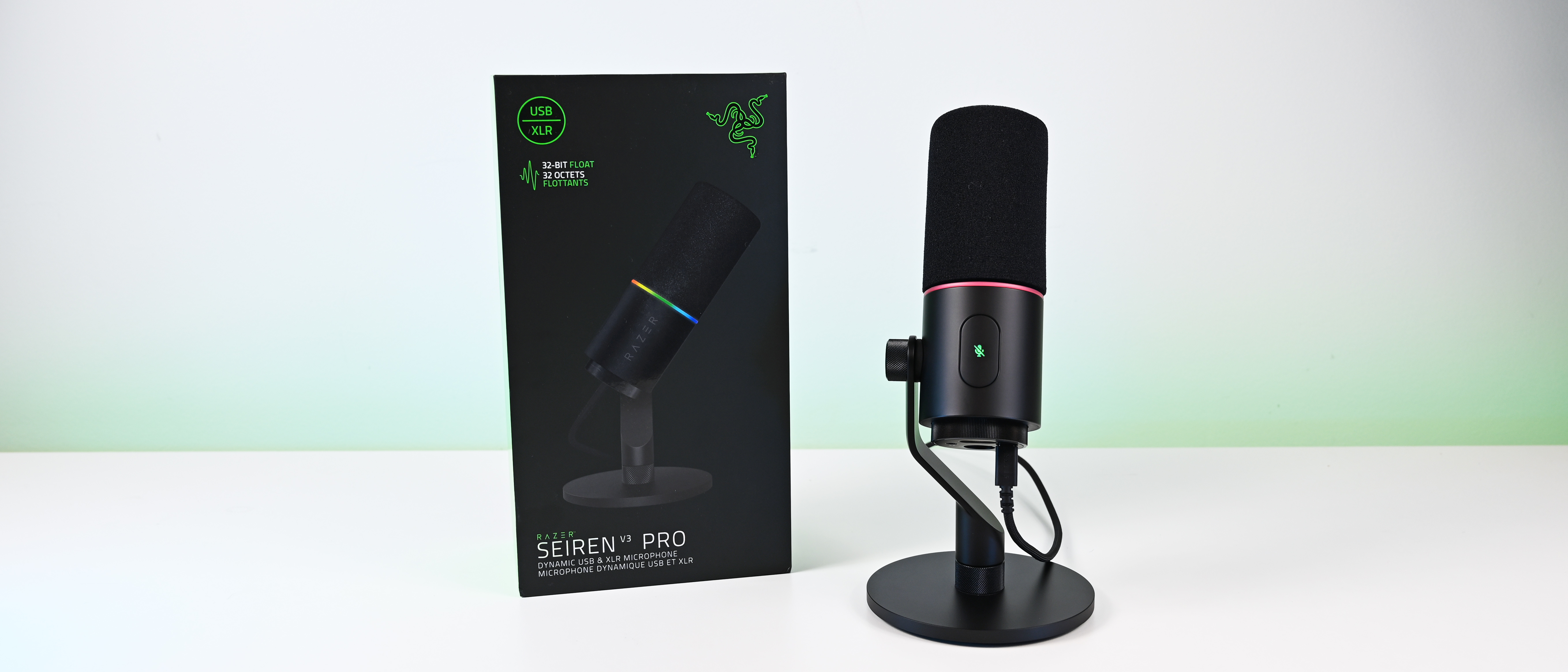 Wide banner shot of the black Razer Seiren V3 Pro retail box standing next to the microphone on its desktop stand on a white table.