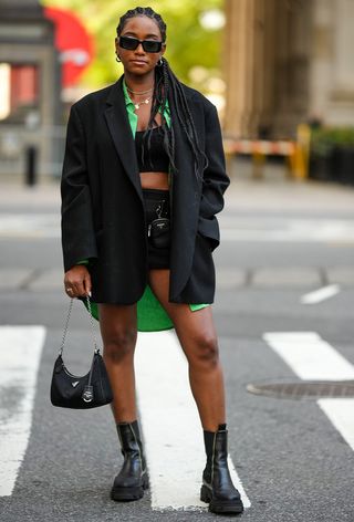 a street-style photo of a woman wearing one of the most popular vintage bags, Prada nylon bag in black styled with a black blazer, green button-down, black bra, black shorts, and black ankle boots