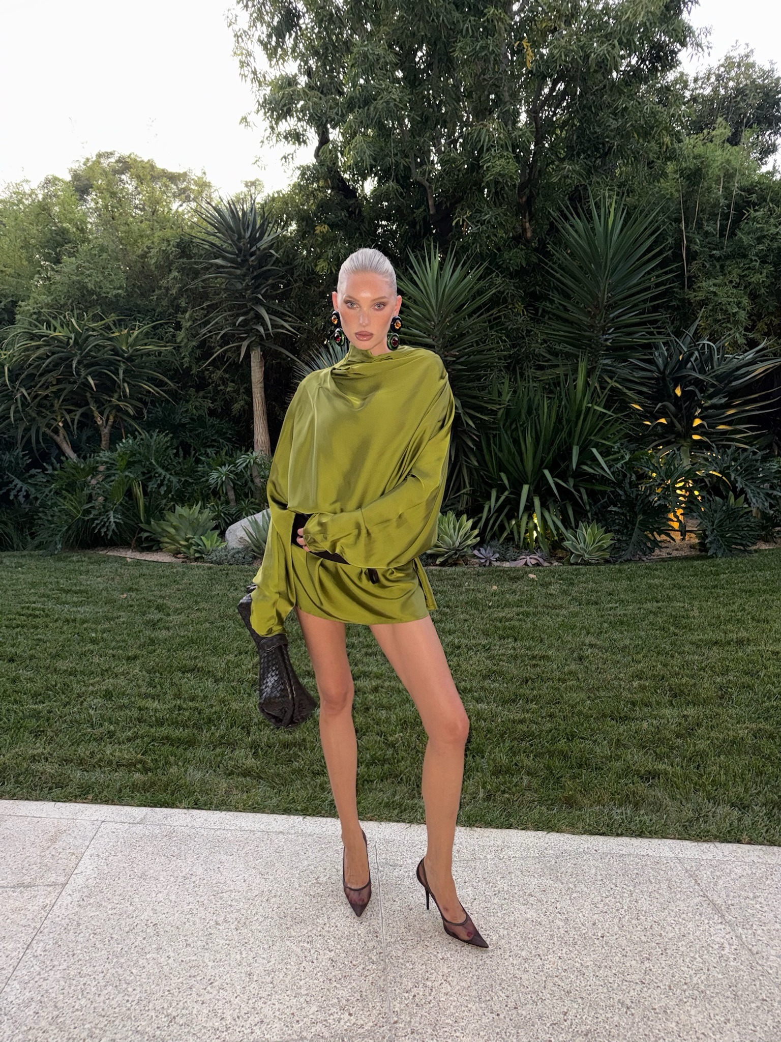 Elsa Hosk wears a green satin dress with a black woven bag and black mesh heels.
