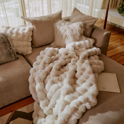 Beige corner sofa piled with white faux fur cushions and throw