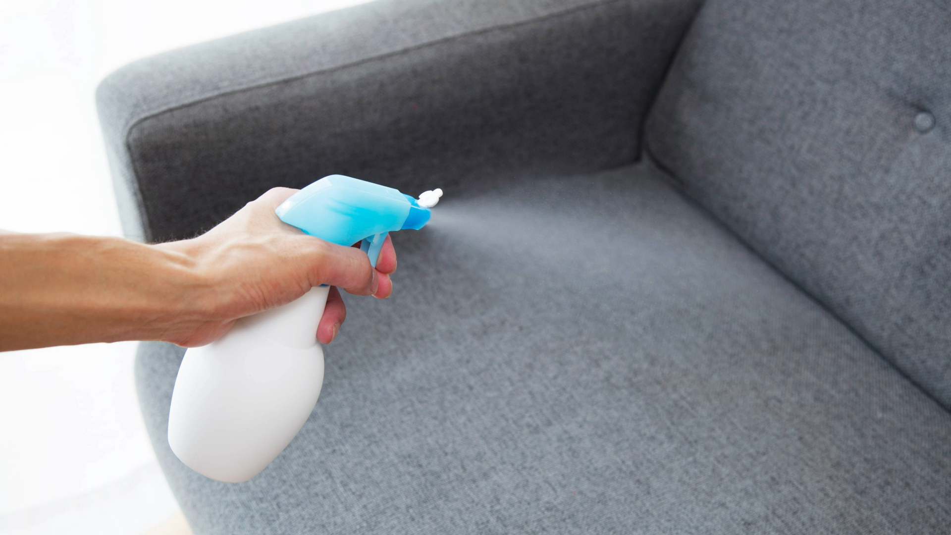 Hand spraying a bottle of liquid on a fabric grey couch