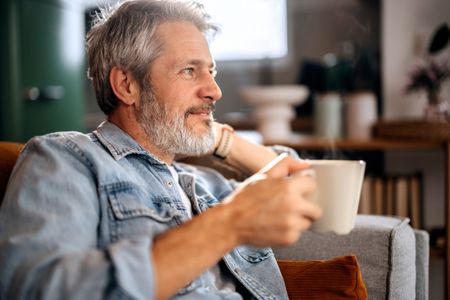 Senior man relaxing on a sofa while drinking coffee at home, calm lifestyle, leisure time and comfortable living concept.