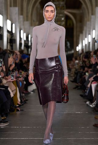 Model walking the Tory Burch fall runway show wearing a purple hooded shirt with a purple leather skirt, tights, and purple pumps.