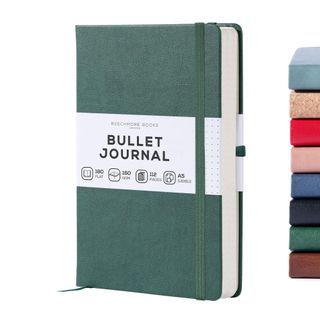 A green bullet journal with a white label with black text and matching elasticated closure next to a stack of eight other journals in various colors and textures