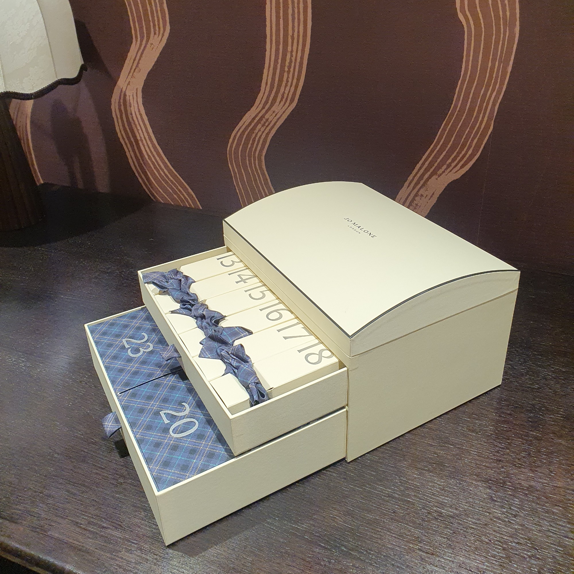 An open Jo Malone luxury advent calendar for 2026 set on a dark wood table with a brown patterned wallpaper in the background