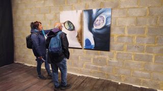Two people with backpacks stand in an art gallery, viewing two large close-up paintings of cat eyes displayed on a gray cinderblock wall.