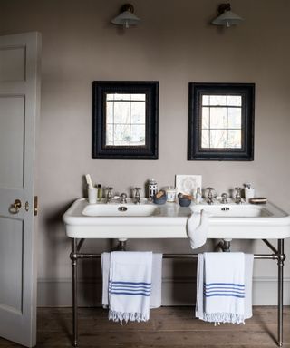 Bathroom with traditional twin basin sink, exposed rails underneath with towels hanging, matching black square mirrors above sink, matching wall lights beside mirrors, dark wood flooring