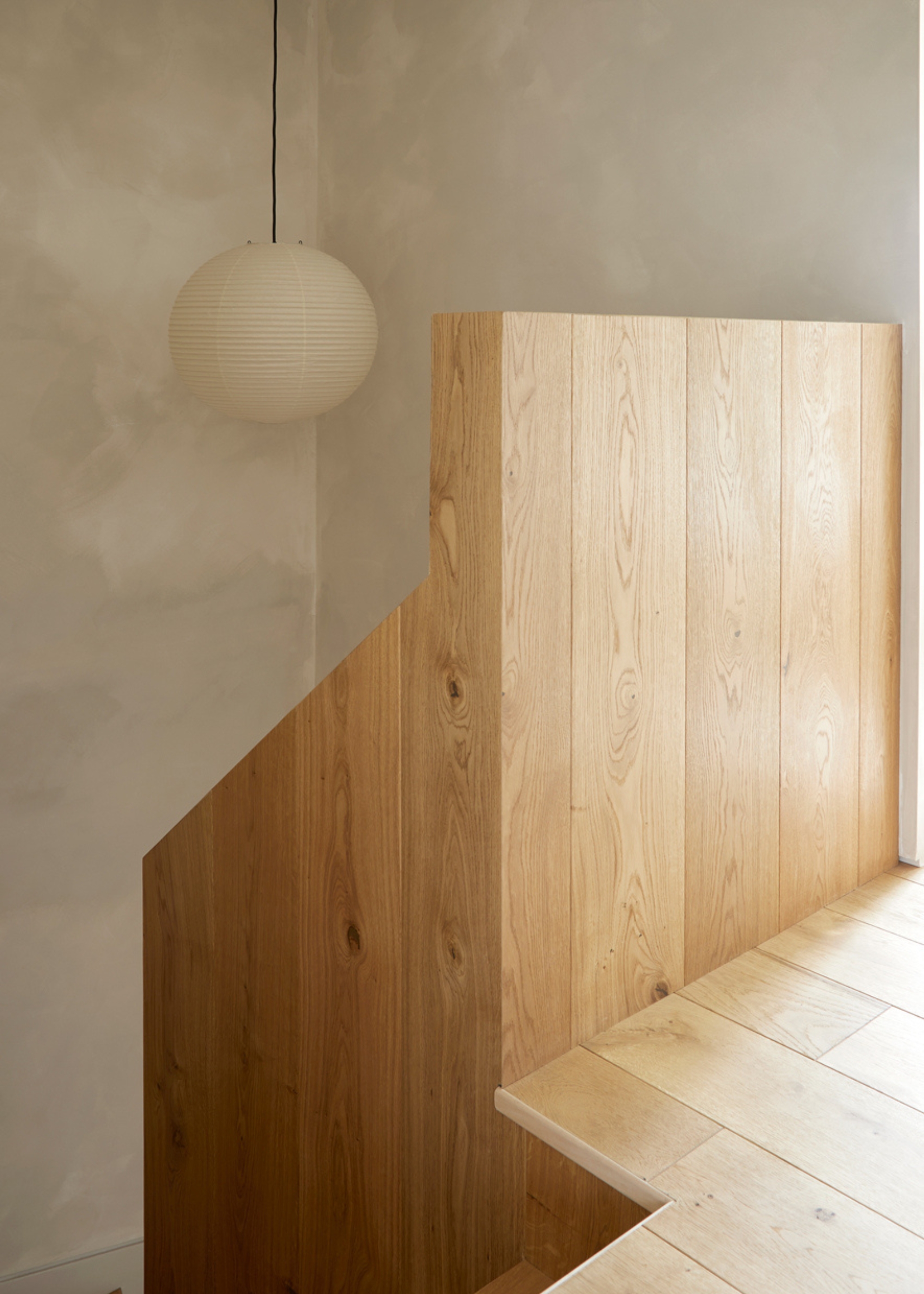 A limewashed wall beside a wood staircase and a rice paper pendant lamp