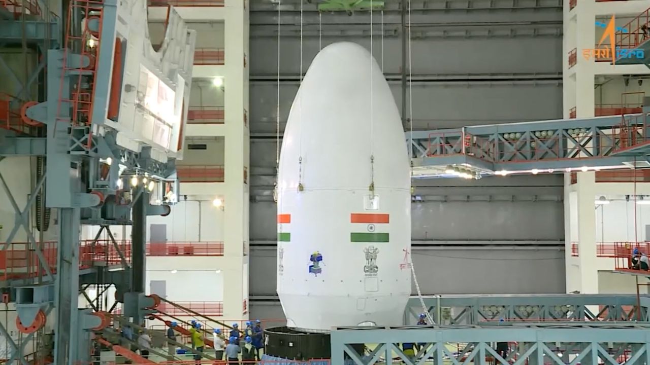 a white conical payload fairing is lowered onto a rocket by wires inside a huge building